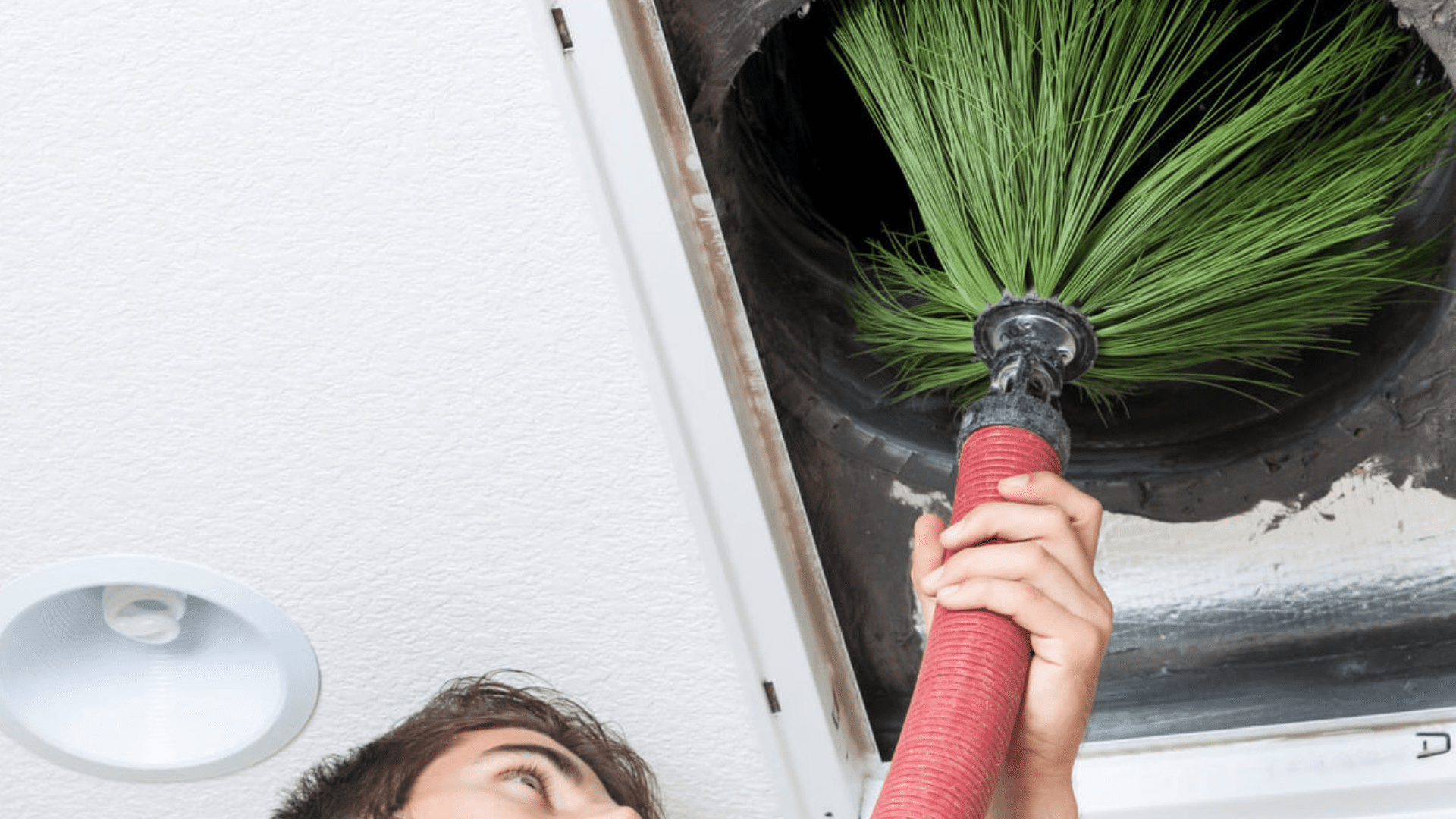 A person using a specialized cleaning tool to remove debris from an air duct in the ceiling, with a focus on the cleaning brush inserted into the duct.