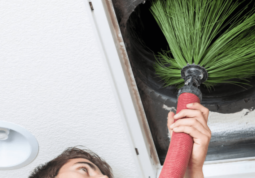 A person using a specialized cleaning tool to remove debris from an air duct in the ceiling, with a focus on the cleaning brush inserted into the duct.